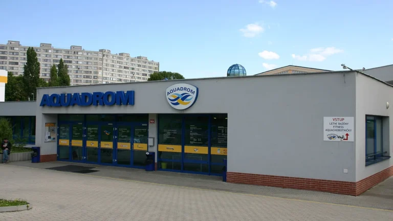 Vstup do přízemní budovy Aquadromu v Mostě s modrým nápisem, v pozadí prosklená kopule a panelové domy