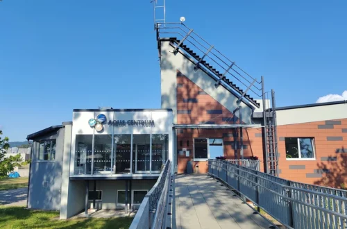 Vstupní budova Aquacentra Šumperk s proskleným vchodem a nápisem Aqua Centrum, k ní vede lávka se zábradlím, za ní věž s cihlovou fasádou