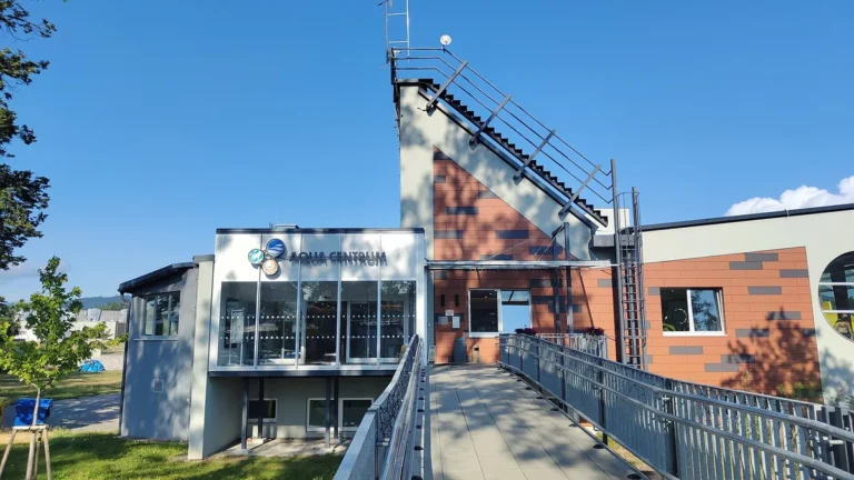 Vstupní budova Aquacentra Šumperk s proskleným vchodem a nápisem Aqua Centrum, k ní vede lávka se zábradlím, za ní věž s cihlovou fasádou