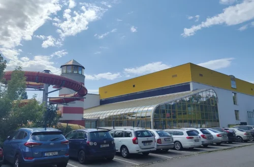 Budova Aquaparku Vyškov se žlutou fasádou a prosklenou halou, vlevo věž v podobě majáku s červeným tobogánem, před ní parkoviště s auty