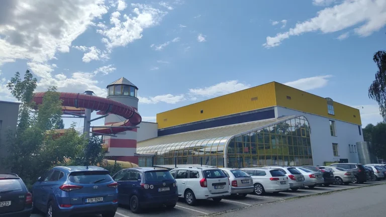 Budova Aquaparku Vyškov se žlutou fasádou a prosklenou halou, vlevo věž v podobě majáku s červeným tobogánem, před ní parkoviště s auty