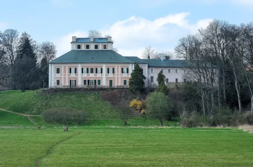 Růžová fasáda zámku Ratibořice nad travnatým svahem, pohled od řeky Úpy v Babiččině údolí
