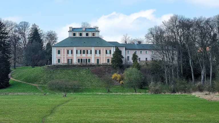 Růžová fasáda zámku Ratibořice nad travnatým svahem, pohled od řeky Úpy v Babiččině údolí