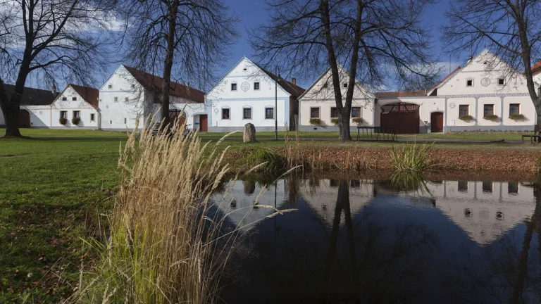 Bílé štíty selských usedlostí na západní straně návsi v Holašovicích odrážející se v návesním rybníčku
