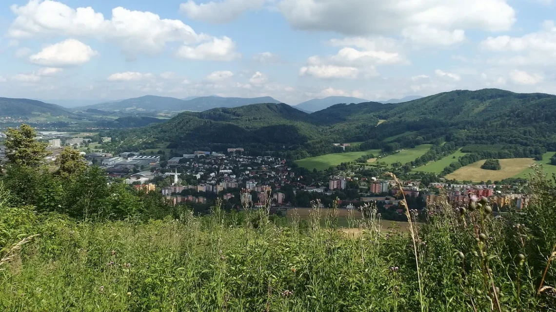 Pohled z kopce na Kopřivnici s panelovými domy a kostelní věží, za městem zalesněné vrchy Beskyd pod oblačnou oblohou