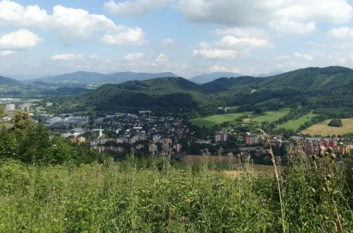 Pohled z kopce na Kopřivnici s panelovými domy a kostelní věží, za městem zalesněné vrchy Beskyd pod oblačnou oblohou
