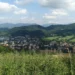 Pohled z kopce na Kopřivnici s panelovými domy a kostelní věží, za městem zalesněné vrchy Beskyd pod oblačnou oblohou