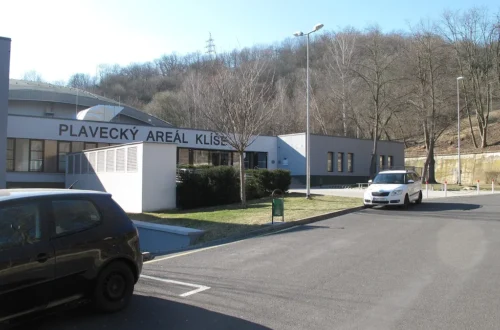 Vchod do přízemní budovy s nápisem Plavecký areál Klíše, před ní parkoviště s auty a v pozadí zalesněný svah