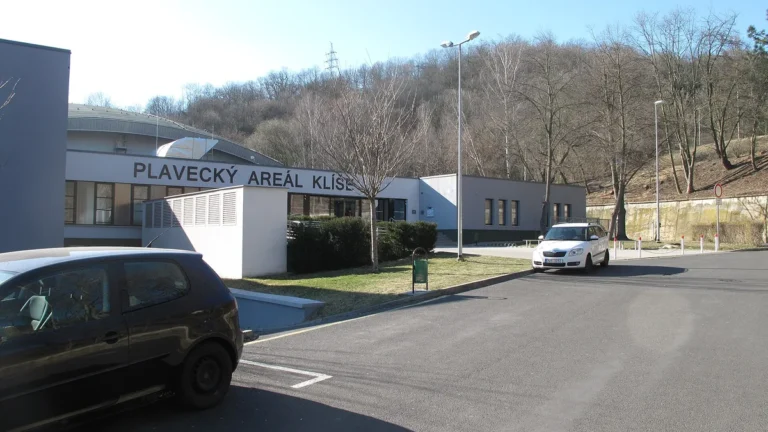 Vchod do přízemní budovy s nápisem Plavecký areál Klíše, před ní parkoviště s auty a v pozadí zalesněný svah