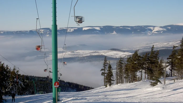 Zasněžená sjezdovka a sedačková lanovka v ski areálu Ramzová s výhledem na hřebeny Jeseníků nad inverzí