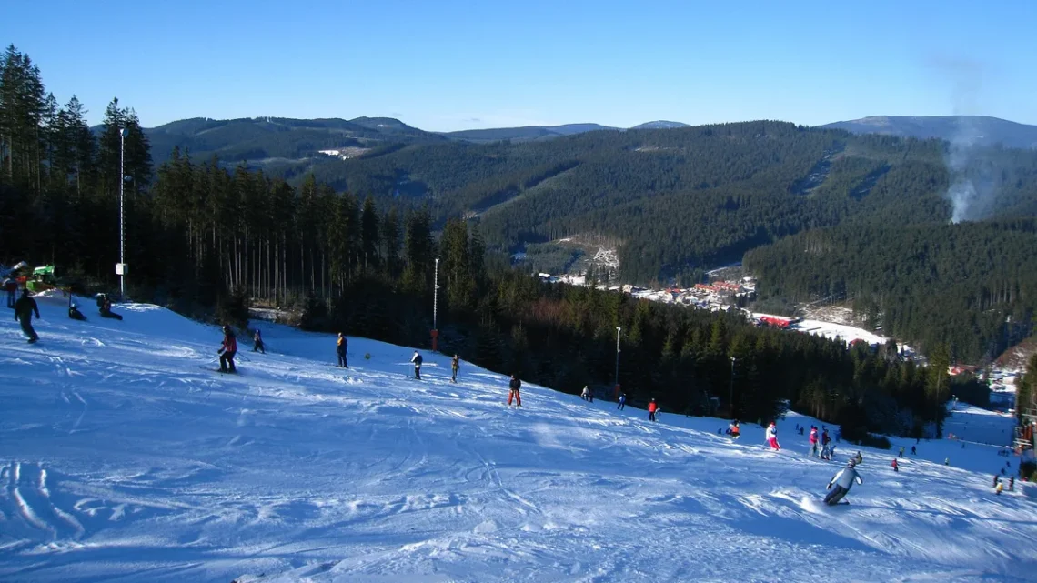 Lyžaři na zasněžené sjezdovce ve skiareálu Bílá s výhledem do zalesněného údolí Beskyd
