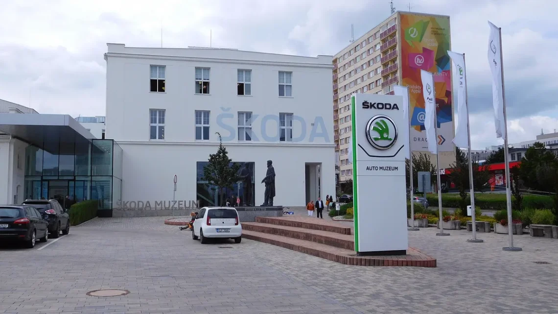 Bílá budova Škoda Muzea na třídě Václava Klementa v Mladé Boleslavi se sochou zakladatelů a stojanem s logem Škoda
