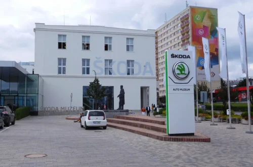 Bílá budova Škoda Muzea na třídě Václava Klementa v Mladé Boleslavi se sochou zakladatelů a stojanem s logem Škoda