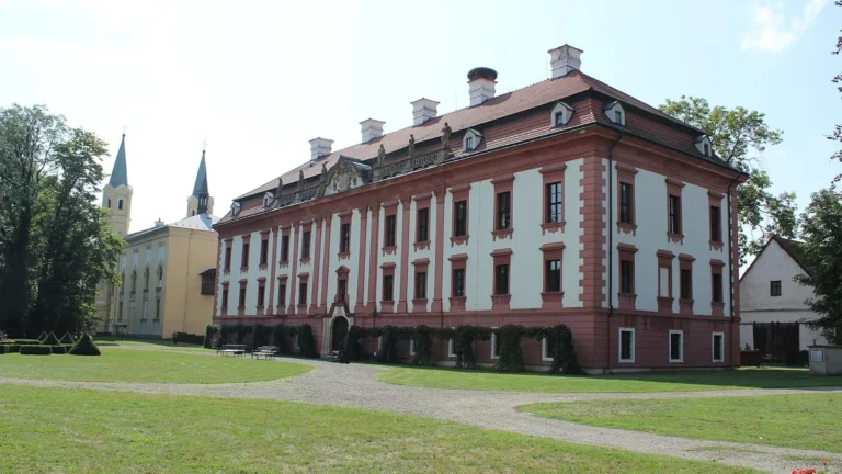 Barokní zámek Kunín s růžovým průčelím, trávníkem zámeckého parku a dvouvěžovým kostelem Povýšení svatého Kříže vlevo