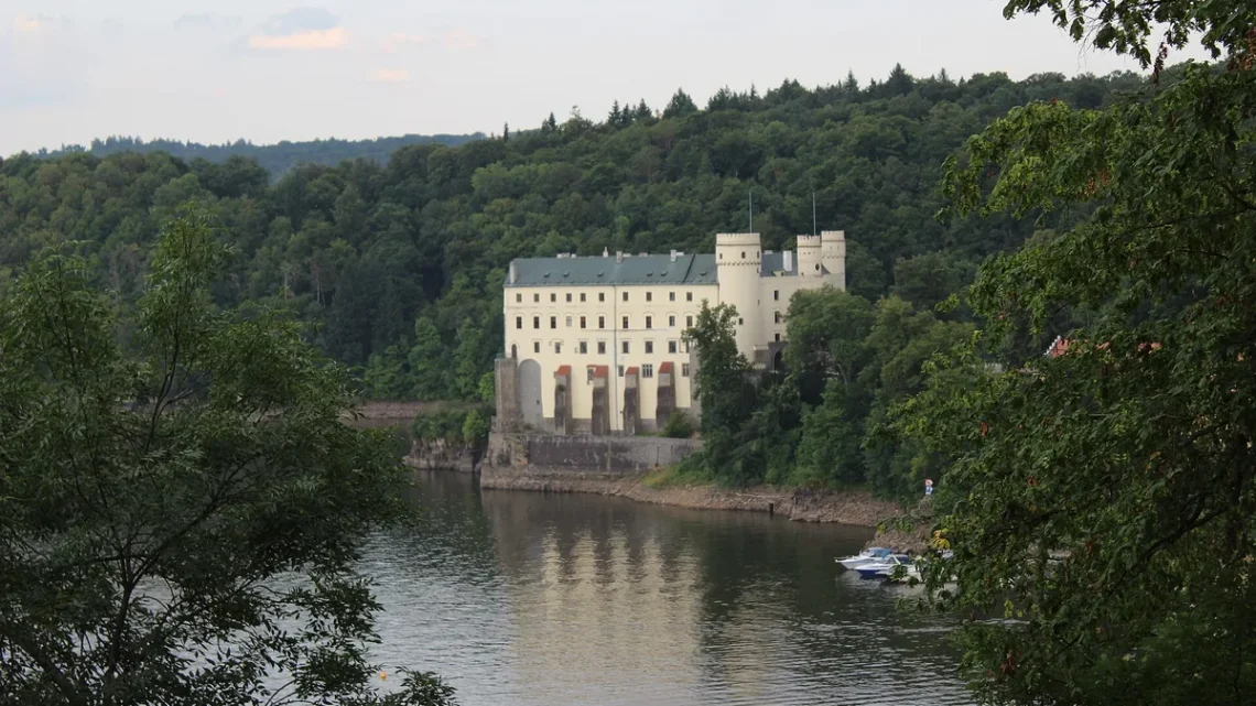 Bílý novogotický zámek Orlík s věžemi nad hladinou Orlické přehrady, obklopený zalesněnými svahy