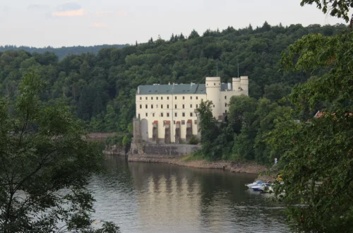 Bílý novogotický zámek Orlík s věžemi nad hladinou Orlické přehrady, obklopený zalesněnými svahy