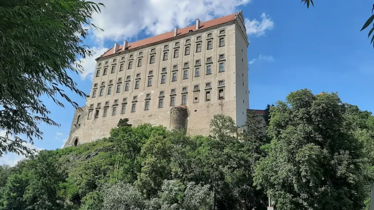 Vysoký a úzký zámek Plumlov na skalnatém návrší nad stromy, pohled zespodu za slunečného dne